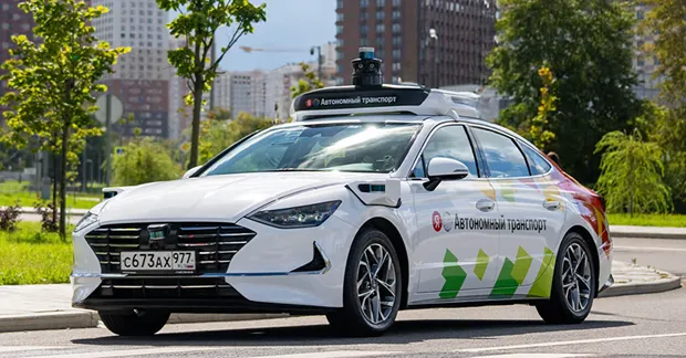 Современный беспилотный автомобиль белого цвета, оборудованный системой автономного управления. На крыше размещён лидар для сканирования окружающей среды. На кузове — надпись Автономный транспорт и яркие графические элементы. Машина движется по городской дороге, на фоне видны многоэтажные здания и деревья