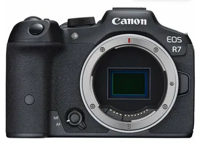 Фронтальный вид беззеркальной камеры Canon EOS R7 с открытым байонетом для установки объектива. Корпус чёрного цвета с текстурированной рукояткой, хорошо виден сенсор камеры. Логотип «Canon» и надпись «EOS R7» расположены на верхней части корпуса