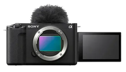 Фронтальный вид камеры Sony Alpha. Корпус выполнен в чёрном цвете, с текстурированной рукояткой слева. Отличительные особенности: откидной экран, развёрнутый вбок, и пушистый ветрозащитный микрофон сверху. Объектив не установлен, виден байонет и матрица